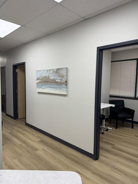 Office hallway with a coastal painting on the wall and a doorway into a small meeting area with two black chairs and a white table.