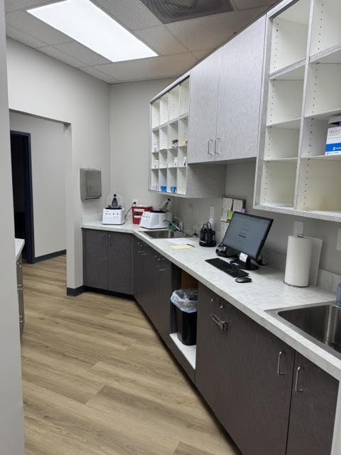Small clinical kitchenette with gray lower cabinets, white countertops, a computer on the right, and open shelving above.